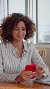 Vertical video of latin business woman in 30s texting on red mobile phone beside laptop at office table, curly haired employee reading text message, filmed as static medium shot of young worker.