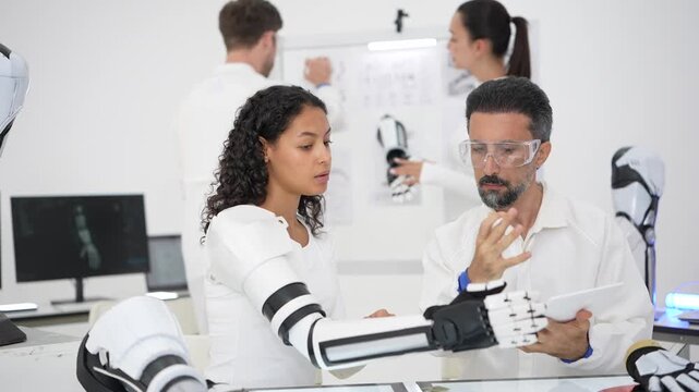 Young woman wearing a full bionic exoskeleton arm raises the robotic hand while a senior scientist inspects a chip, team working behind
