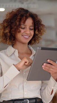 Vertical video of curly haired business woman in 30s checking digital report on tablet touchscreen inside office, multiracial employee reading online data, medium shot of corporate company staff.