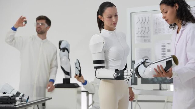 Woman wearing an exoskeleton arm examines the robotic hand while an engineer holds up a microchip, prosthetic research in a white lab