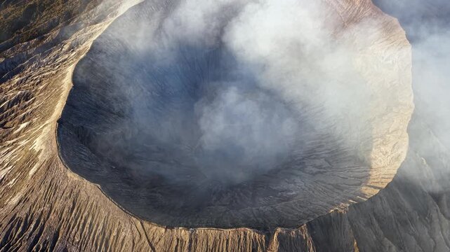 Sunrise drone view of Mount Bromo crater rim with steam and golden light illuminating rugged volcanic landscape.