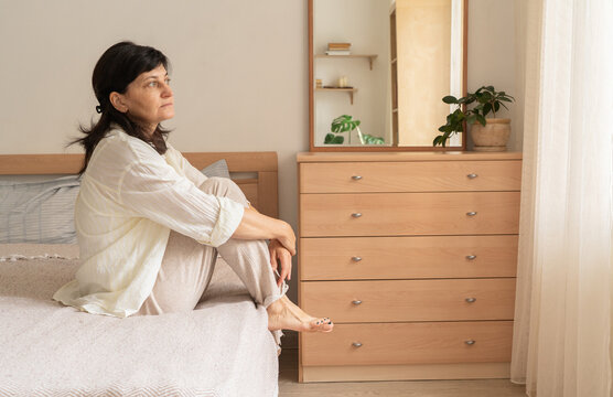 Woman sits on bed with her knees drawn up to her chest, gazing out bedroom window. She is deep in thought. Women's mental health, slow mindful morning