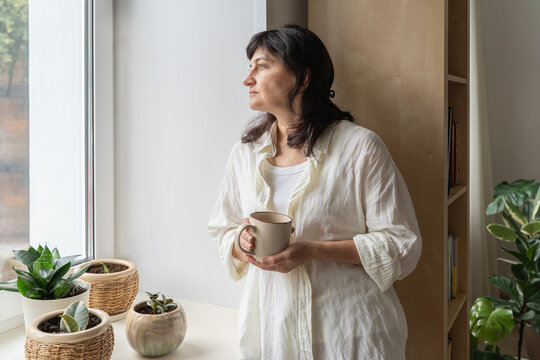 Mature woman gazes thoughtfully out window, holding cup of hot beverage, cozy home setting. A slow paced home life, mindful living after 50, and womens mental and hormonal health