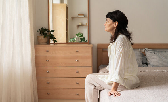 Mature woman in loungewear sits on edge of bed in bright bedroom, gazing out the window with pensive and sad expression, suggesting depression or mental health issues