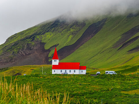 Green fields and Vik i Myrdal Lutheran church on the hill. White building with a red roof and tower. In the background, visible M&yacute;rdalssandur mountain slope is covered with rain clouds. Vik, Iceland.