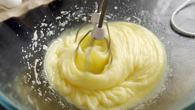 Mixing eggs and sugar in a glass bowl with electric mixer, creating a smooth batter, eggshells scattered on the counter