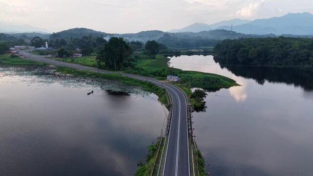 Aerial winding asphalt road flanked by reflective water bodies and surrounded by lush green vegetation under a partly cloudy sky in a serene natural setting at Tasik Raban