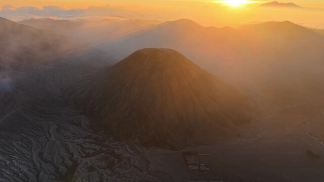 Drone aerial view of Gunung Batok dark volcanic cone, rugged ash terrain, distant mountains, and glowing sunset haze.