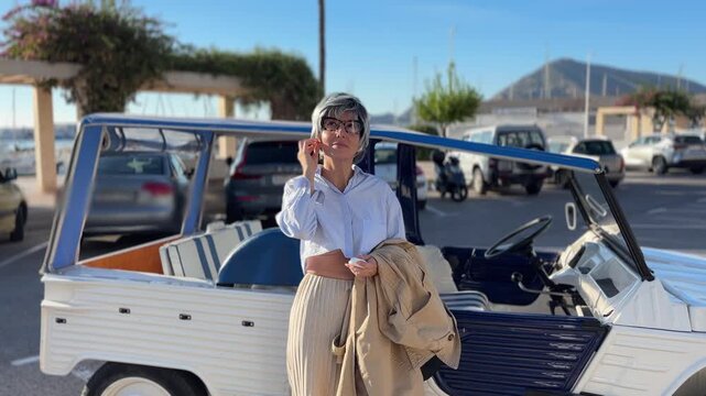 Mujer +50 poni&eacute;ndose auriculares junto a coche cl&aacute;sico en el puerto