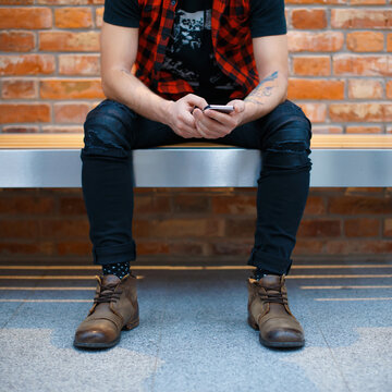 Stylish young man in plaid shirt sitting on a bench against a brick wall, holding and using a smartphone. Concept of urban lifestyle, modern communication, texting, and mobile technology.