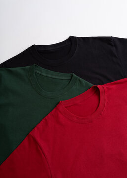 Top view of three folded blank crew neck t-shirts in black dark green and red colors arranged on a white background