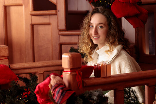 The woman with curly hair wears a soft fleece jacket while resting on a rustic wooden staircase adorned with festive holiday greenery and ornaments