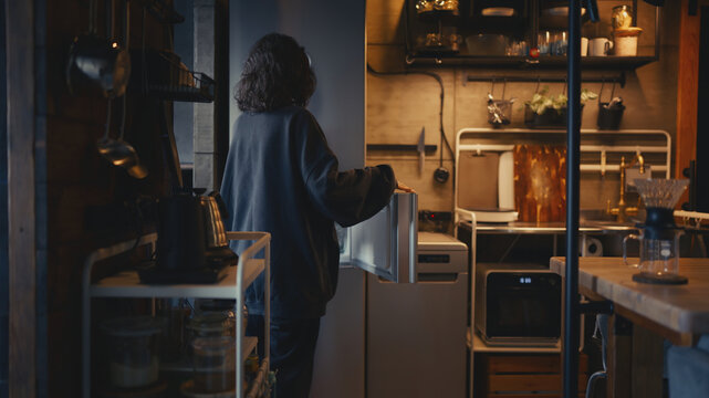 Candid back view of a woman in cozy, warm-lit evening loft kitchen with open shelves opening the fridge. Concept of healthy dinner preparation, modern domestic lifestyle, daily routines, food choice. 