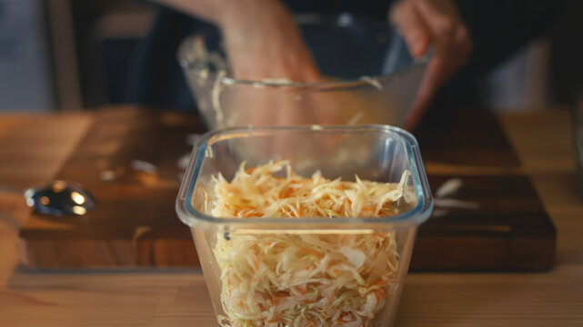 Close-up of a glass container filled with seasoned shredded cabbage and carrots. Woman's hands preparing homemade fermented sauerkraut in a bowl on a wooden table in warm home kitchen.