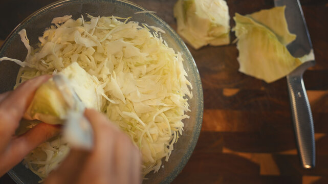 Top view close-up of woman's hands shredding fresh cabbage with a peeler into a full glass bowl on a wooden cutting board. Cooking and healthy food preparation concept in a home kitchen. 