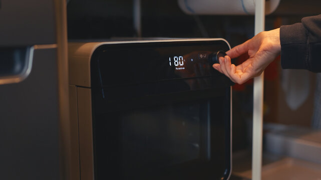 Close-up of a woman's hand turning a knob on a portable electric mini oven to adjust preheat mode temperature in a cozy kitchen. Preparing to bake or roast food at home.