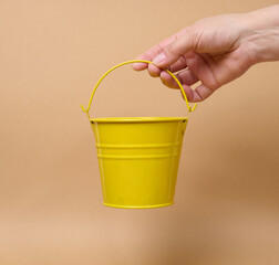 A person holding a small yellow bucket by the handle against a beige background. © nndanko