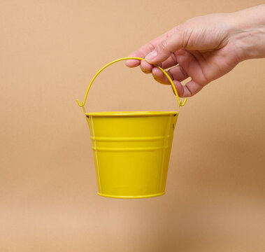 A person holding a small yellow bucket by the handle against a beige background.