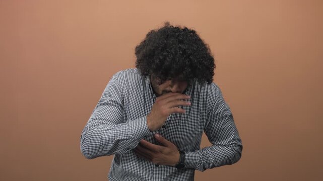 Man clutching chest with hands coughing and grimacing in studio with peach wall, curly hair visible; pain heartattack.
