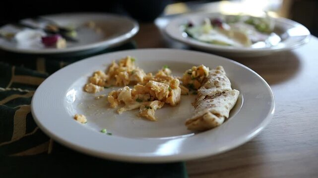 Detailed view of a delicious hot breakfast served on a white plate in a restaurant, featuring scrambled eggs or omelet with a wrap roll on a dining table.