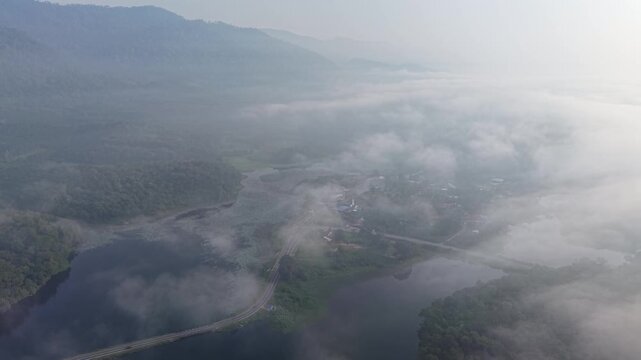 Aerial view captures misty mountain peaks. Fog drifts over green valley fields at Tasik Raban