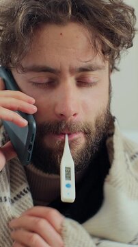 Vertical video. Portrait of sick man sitting in room and talking on the phone while sitting on the sofa. Ill infected male coughing and having pain in chests at home while talking on smartphone.