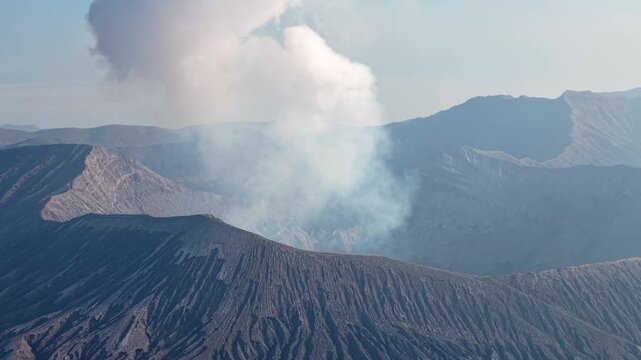White smoke rises from a rugged mountain peak. Steam drifts across dark, craggy volcanic slopes at Mount Bromo