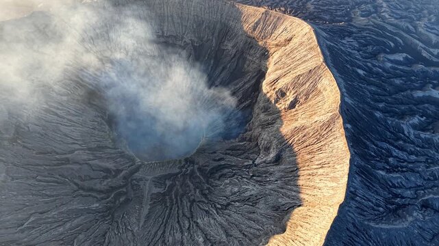 Active volcano Mount Bromo erupts, smoke billows from crater. Rugged cliffs surround deep, dark crater