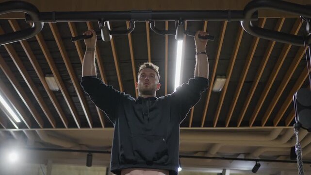 Dynamic low angle slow motion view circles an athletic man performing wide grip typewriter pull ups and moving side to side beneath linear lights in a modern gym.