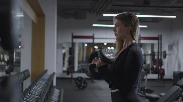 Medium side view of a focused woman completing standing dumbbell biceps curls beside a long free weight rack in slow motion.