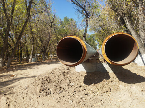 Two large insulated steel pipeline pipes laid on concrete supports in an outdoor autumn park, showing their large open ends against a backdrop of trees and a sunny sky.