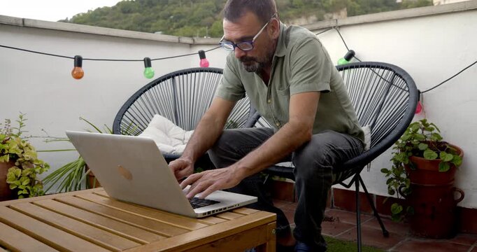 Man Working on Laptop on Rooftop Terrace: A focused man wearing glasses sits on a modern chair on a rooftop terrace, typing on his laptop, with colorful string lights in the background.