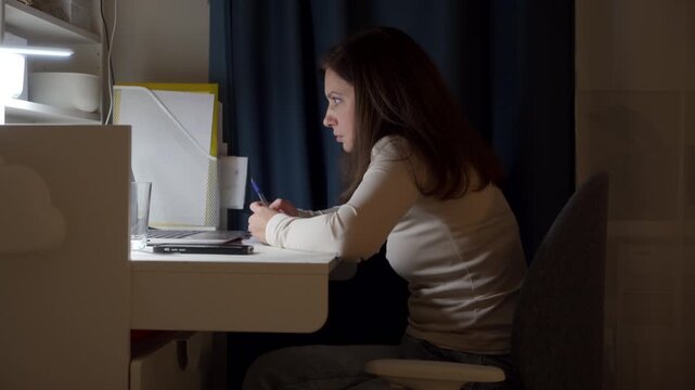 A woman holds a pen while reading her laptop screen. She sits attentively at a white desk in a dark room. Her quiet stance reflects focus and devotion.