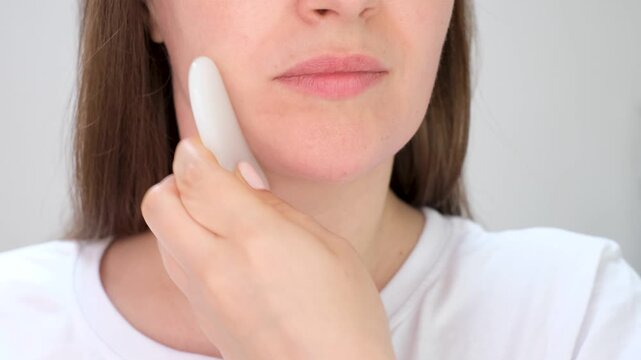 Young woman doing facial massage with massager gua sha on gray background front view.