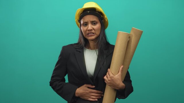 Woman architect, young brunette hispanic in yellow hardhat and blazer, holding rolled blueprints and holding stomach while grimacing in studio; discomfort.