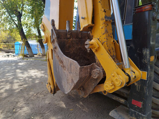 Detailed close-up of a yellow backhoe excavator bucket with steel teeth and a shiny hydraulic cylinder parked on dirt at an outdoor construction site with trees in background. © z_olga_art