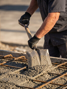 A construction worker spreads wet concrete over steel reinforcement with a shovel. The hands-on process illustrates foundation work, structural development and skilled building labor