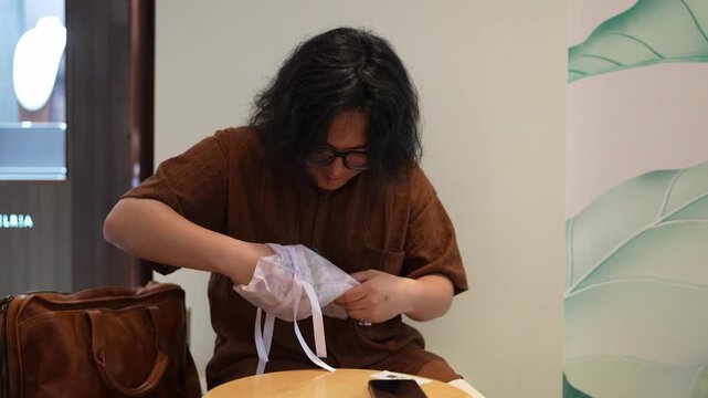 A seated man opens a translucent drawstring pouch while a leather bag rests beside him in a cafe.