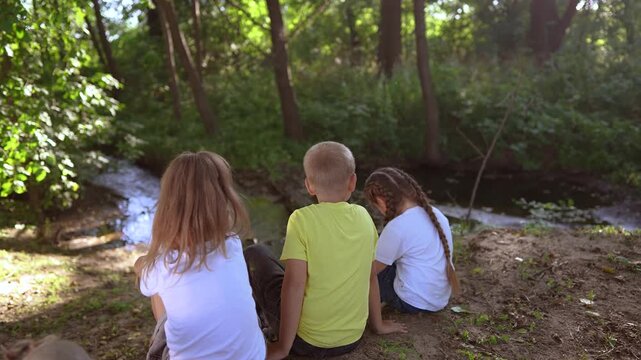 children sit on the banks of river. happy family lifestyle childhood dream concept. a group of little children sit together by the river and talk. kids spend time outdoors in nature in the forest
