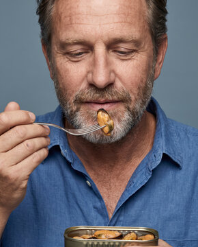 Weathered man eats preserved fish from a tin, with a fork against a solid studio background