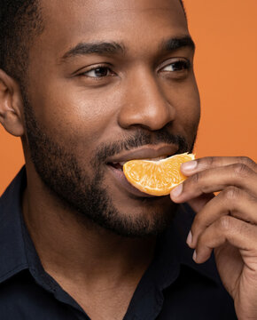 Bright man eats a fresh orange segment, with a gentle smile against a bold color backdrop