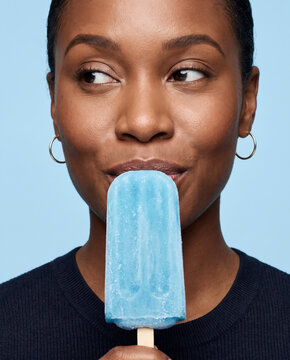 Cool woman eats a blue popsicle, with a sidelong glance in soft beauty lighting