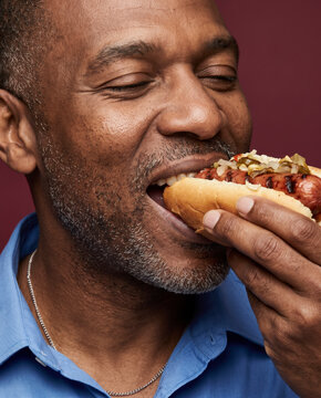 Hearty man bites a loaded hot dog, with relish with striking studio color