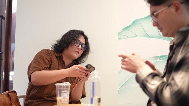 A seated man smiles and looks down while another man gestures beside the cafe table.