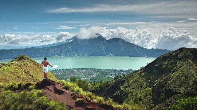 Hiker stands on Mount Batur Caldera Ridge, taking off his shirt after hard hiking, enjoying Lake Batur and the lush volcanic landscape, adventure and exploration in Bali. Aerial view drone flight