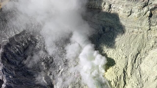 Volcanic smoke rises from a cratered peak. Steam swirls over rugged, mineral stained rock faces at Mount Ijen