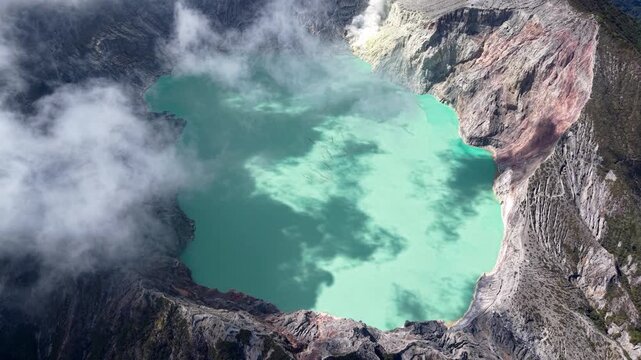 Aerial drone over Mount Ijen turquoise crater lake with steaming fumaroles and rugged volcanic cliffs.