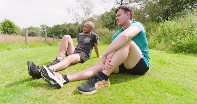 Two men resting on grass after jogging, enjoying conversation in park