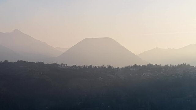 Misty mountains glow at dawn. Silhouettes frame distant peaks. Village lies nestled in shadows at Bromo