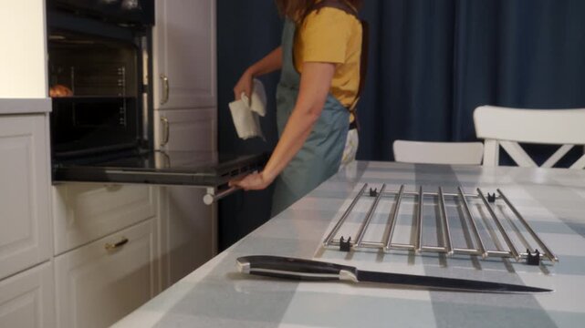 A woman in an apron opens a warm oven in a modern kitchen. She holds a towel while preparing to remove hot food. Cooking at home brings cozy feelings of comfort and quiet joy.
